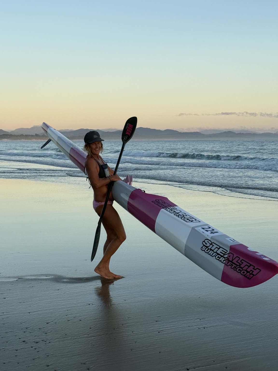 Heli smiling on the beach while carrying her ocean ski and paddle