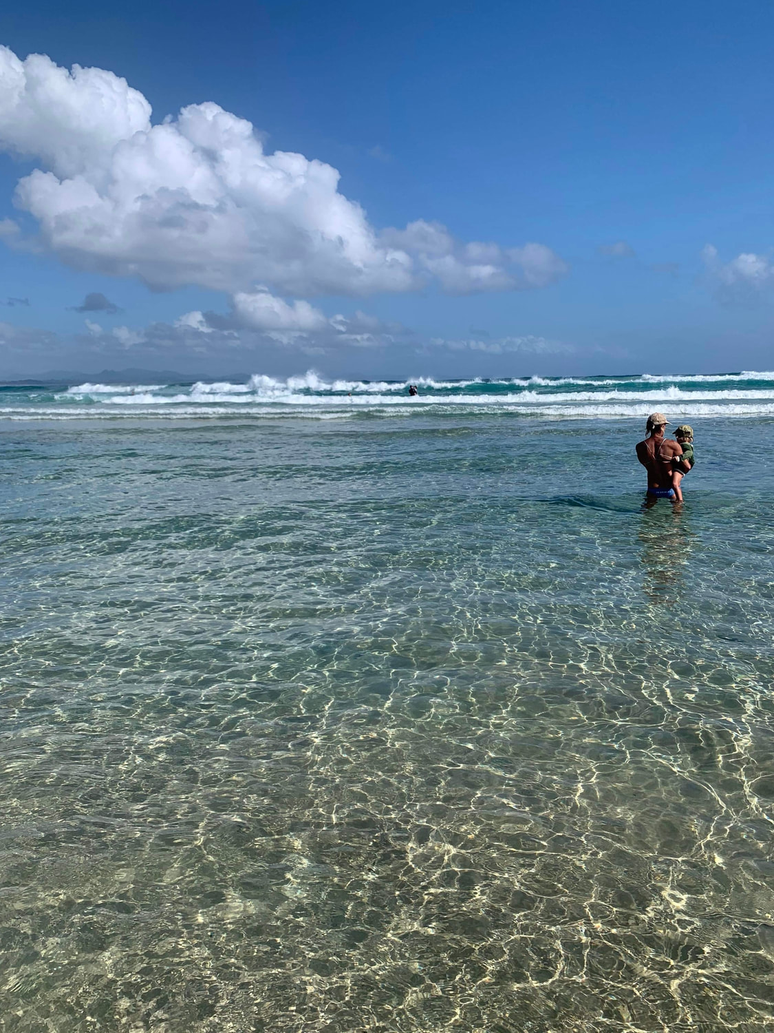 Heli holding her grandson in clear ocean water beneath a wide blue sky
